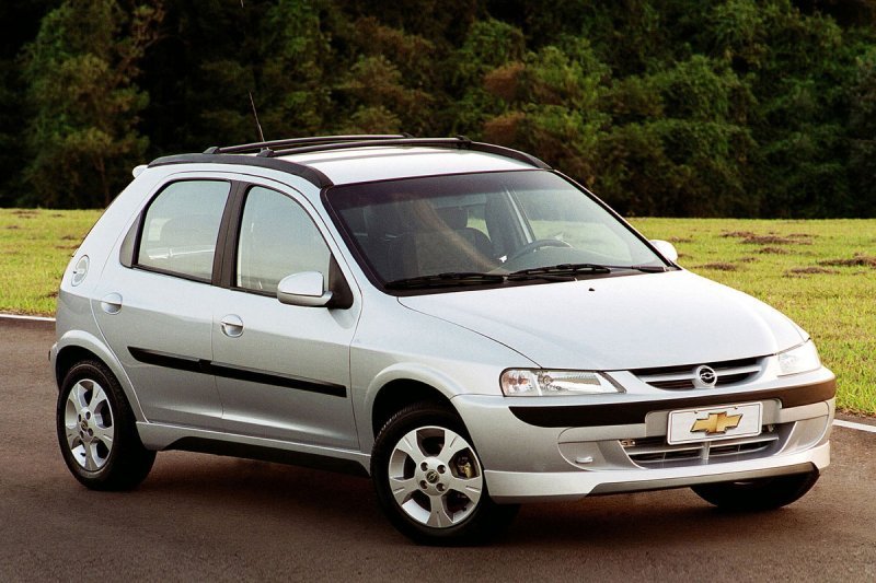Chevrolet Celta Super prata 4 portas da geração antiga estacionado em via asfaltada com vegetação ao fundo.