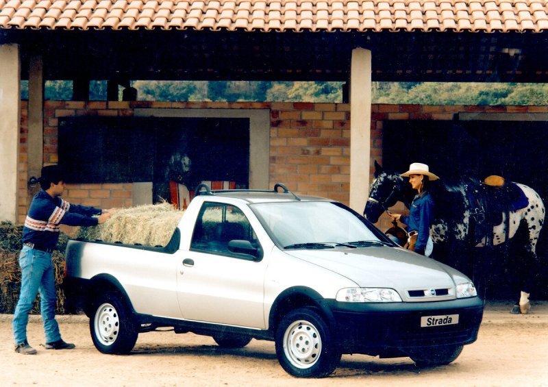 Fiat Strada 1ª geração sendo carregada com feno em fazenda, ao lado de cavalo e vaqueiros. Utilizada para trabalho rural.