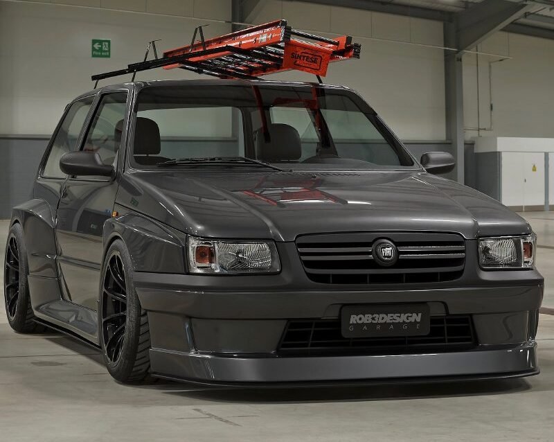 Fiat Uno escada rebaixado cinza com bodykit widebody, rodas pretas e escada laranja no teto em pavilhão