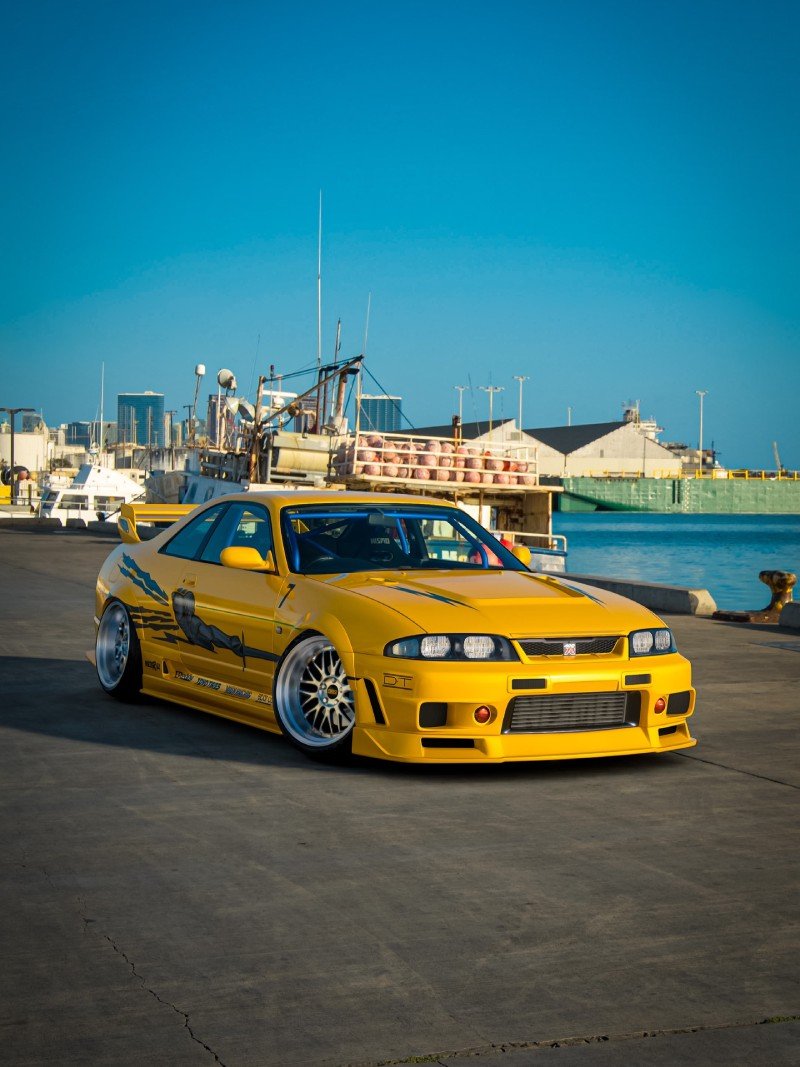Nissan Skyline GT-R R33 amarelo rebaixado em foto 3/4 dianteira, com rodas BBS e fundo de porto